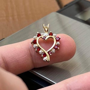 May include: A gold-colored heart-shaped pendant with a halo of red and clear gemstones. The pendant features a delicate open heart design, with a small loop at the top for a chain. The gemstones are round-cut and set closely together.