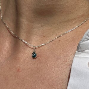 Puede incluir: Un collar de cadena de plata con un peque&ntilde;o colgante azul de ojo turco. El collar lo lleva una persona con una camisa blanca.