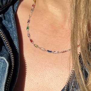 May include: A silver chain necklace with rectangular multi-colored gemstones. The necklace is on a person's neck.