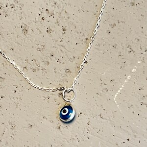 Puede incluir: Un collar de cadena de plata con un peque&ntilde;o amuleto azul en forma de ojo.
