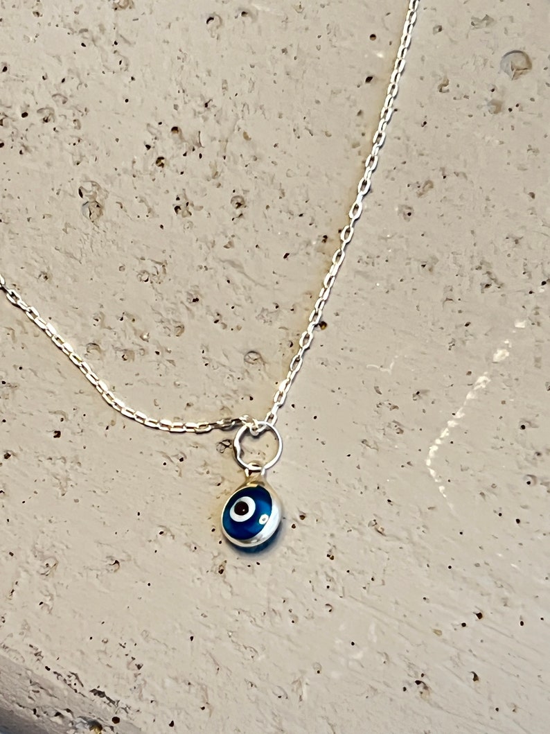 Puede incluir: Un collar de cadena de plata con un peque&ntilde;o amuleto azul en forma de ojo.
