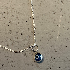 Puede incluir: Un collar de cadena de plata con un dije de ojo turco azul. El dije es redondo y tiene un centro blanco.