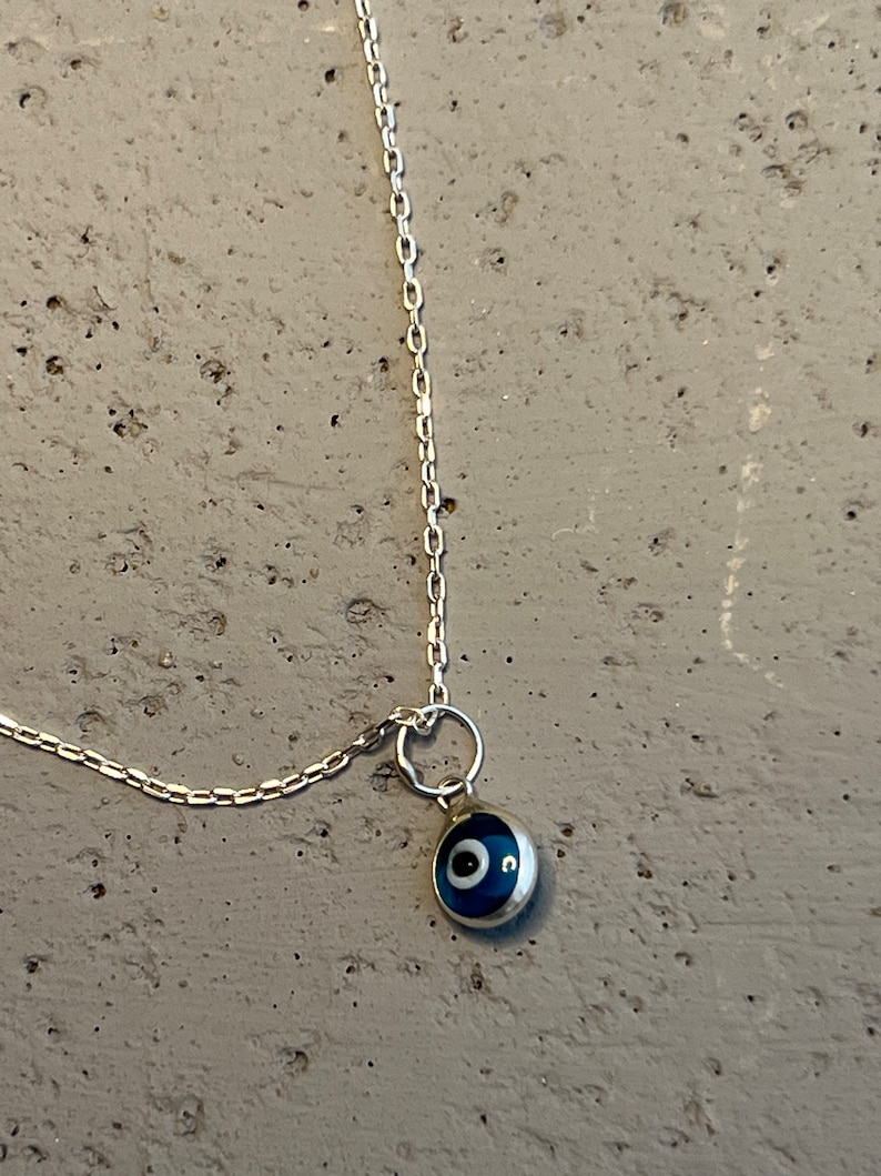 Puede incluir: Un collar de cadena de plata con un dije de ojo turco azul. El dije es redondo y tiene un centro blanco.
