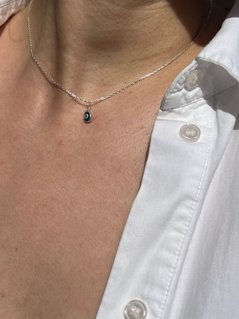 Puede incluir: Un collar de cadena de plata con un peque&ntilde;o colgante azul de ojo turco. El collar lo lleva una persona con una camisa blanca.