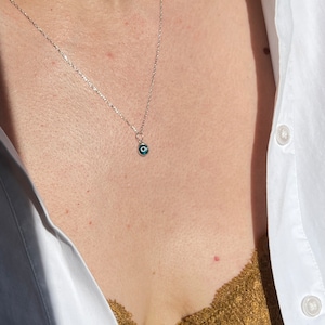 Puede incluir: Un collar de cadena de plata con un peque&ntilde;o dije azul de ojo turco. El collar lo lleva una persona con una camisa blanca y un top de encaje marr&oacute;n.