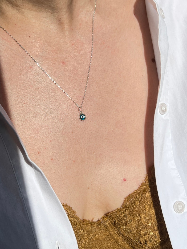Puede incluir: Un collar de cadena de plata con un peque&ntilde;o dije azul de ojo turco. El collar lo lleva una persona con una camisa blanca y un top de encaje marr&oacute;n.
