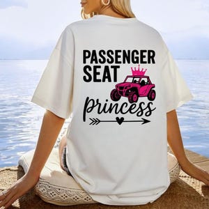 May include: Cream-colored t-shirt with the words "PASSENGER SEAT princess" in black script and a pink off-road vehicle graphic. The design includes a crown and an arrow with a heart.