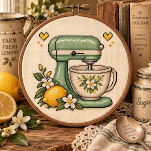 Op de afbeelding: Een ronde houten borduurring met een kruissteekpatroon van een lichtgroene keukenmixer met een witte mengkom versierd met witte bloemen. Een gele citroen en witte bloesems zijn aan de voet van de mixer geplaatst.