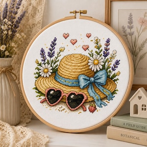 May include: A cross-stitch embroidery in a wooden hoop featuring a tan straw hat with a blue ribbon, heart-shaped pink sunglasses, and a floral arrangement of white daisies and purple lavender sprigs, accented by small pink hearts and a strawberry.