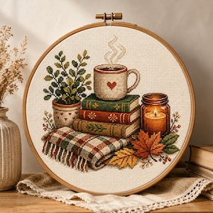 Puede incluir: Un bastidor de bordado de punto de cruz con una escena otoñal. Representa una pila de libros, una taza humeante con un corazón, una vela encendida, una planta en maceta y una manta a cuadros. La paleta de colores incluye marrones, verdes y naranjas cálidos.
