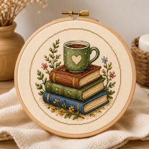 Könnte beinhalten: Ein runder Stickrahmen aus Holz mit einem Kreuzstichmotiv, das eine grüne Tasse mit einem weißen Herz auf einem Stapel aus drei braunen, grünen und blauen Büchern zeigt, umgeben von kleinen bunten Blumen und grünem Blattwerk.