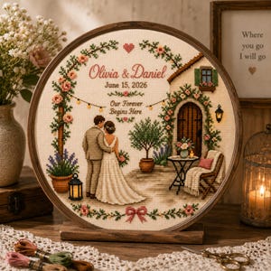 Può includere: Un ricamo a punto croce circolare in un telaio di legno raffigurante una coppia davanti a un edificio in pietra con un arco floreale. Il testo recita Olivia & Daniel, June 15, 2026, e Our Forever Begins Here, circondato da piante e lanterne.