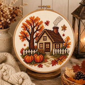 May include: A circular wooden embroidery hoop containing a cross-stitch pattern of a small house with a brown roof, a chimney with smoke, a large orange tree, a white picket fence, and two orange pumpkins on a cream-colored fabric background.