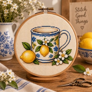 Pode incluir: Um bastidor de bordado circular de madeira contendo um padrão de ponto cruz de uma caneca branca decorada com motivos azuis, limões amarelos e flores brancas. O design inclui um limão amarelo e um cacho de flores brancas com folhas verdes.