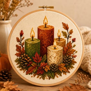 May include: A cross-stitch embroidery in a circular wooden hoop featuring four lit candles in shades of green, orange, beige, and white, surrounded by a decorative arrangement of autumn leaves and pinecones on a cream-colored fabric background.