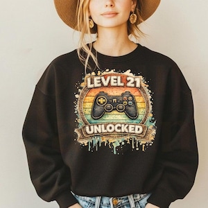 May include: Black sweatshirt with a retro-style graphic. The graphic features a video game controller and the text "LEVEL 21 UNLOCKED" in a circular design with a sunset color scheme. The sweatshirt is a crew neck style.