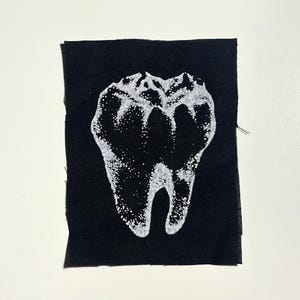 May include: A black fabric patch featuring a white, detailed illustration of a molar tooth. The tooth design is created with small, speckled dots, giving it a textured appearance. The patch has a raw, unfinished edge.