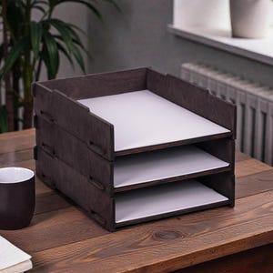 May include: A three-tiered wooden desk organiser in dark brown. Each tier holds a white sheet of paper. A dark brown mug and a notebook are on the wooden desk. A plant is visible in the background.