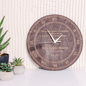 Puede incluir: Un reloj redondo de madera con números romanos y una cita grabada. Las manecillas del reloj señalan aproximadamente la 1:50. El reloj está personalizado con un nombre, universidad y fecha. Hay plantas decorativas en el fondo.