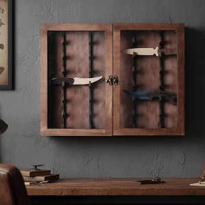 May include: A wooden display cabinet with two glass doors, showcasing knives. The cabinet has a dark brown finish and is mounted on a gray wall. Inside, knives are displayed against a brown backing. The cabinet is closed with a decorative metal clasp.