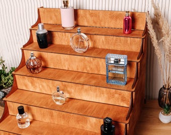 Könnte beinhalten: Ein fünfstufiger Präsentationsständer aus Holz mit einer hellbraunen Holzmaserung. Auf den abgestuften Regalen stehen verschiedene Parfümflakons aus Glas in unterschiedlichen Formen, Größen und Farben, darunter transparente, rosa, blaue und schwarze Glasbehälter.