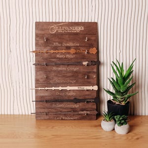Könnte beinhalten: Eine Holztafel mit sechs Zauberstäben, die jeweils mit dem Namen einer Figur aus einer Fantasy-Serie beschriftet sind. Die Zauberstäbe sind in verschiedenen Farben und Designs gehalten. Die Tafel trägt oben den Text "Ollivander's".