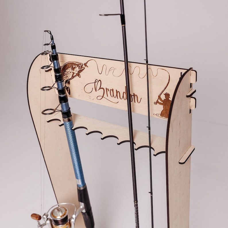 May include: A wooden fishing rod holder with a personalized name, "Brandon", engraved on the front. The holder features a fish and fisherman design. A blue and black fishing rod is in the holder.