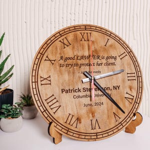 Puede incluir: Un reloj de madera con números romanos y una cita: "A good LAWYER is going to try to protect her client." El reloj también incluye un nombre, una universidad y la fecha de junio de 2024. El reloj está sobre un soporte de madera.