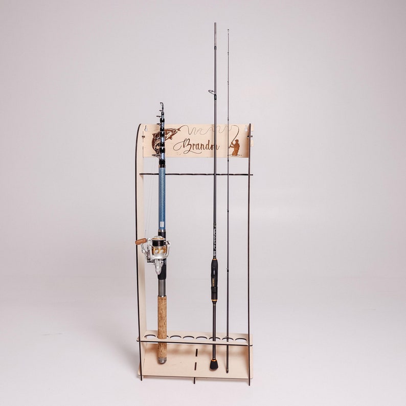 May include: A wooden fishing rod rack with three fishing rods. The rack has a decorative top with the name "Brandon" and a fishing-themed silhouette. The fishing rods are black and blue, and the rack is made of light-colored wood.