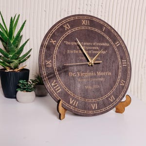 Puede incluir: Un reloj redondo de madera con números romanos y manecillas doradas. El reloj presenta una cita grabada y el nombre "Dr. Virginia Morris" con "Harvard University" y "Mayo de 2024". El reloj se muestra sobre un soporte de madera.