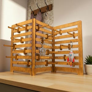 Puede incluir: Un expositor de madera de color marrón claro con diseño plegable, que cuenta con múltiples listones horizontales y clavijas salientes para colgar objetos. Dos llaveros, uno con un dije de brújula circular y otro con un cohete, cuelgan de las clavijas.