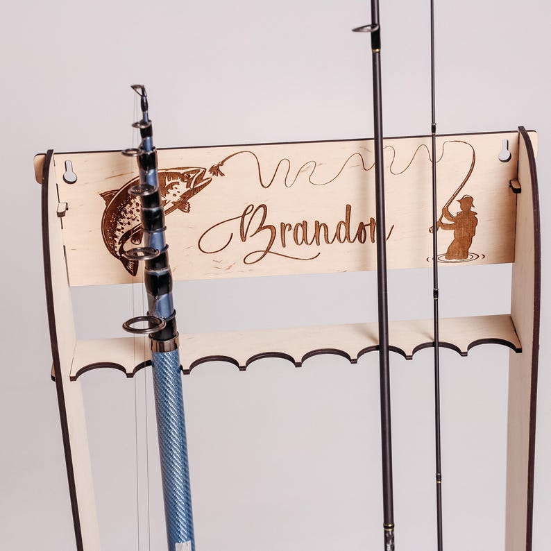 May include: Wooden fishing rod rack with a laser-etched design. The rack features a fish, a fisherman, and the name "Brandon". A blue and black fishing rod is displayed in the rack.