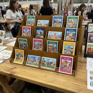 Op de afbeelding: Een bruine, getrapte kartonnen displaystandaard met meerdere rechthoekige ansichtkaarten met kleurrijke illustraties van Oekraïense steden. Elke kaart toont een stadsnaam zoals Kyiv, Lviv en Chernivtsi, met levendige architecturale ontwerpen.