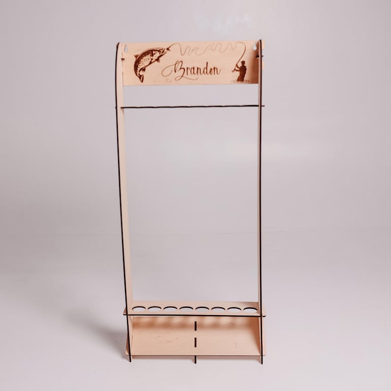 May include: A wooden fishing rod rack with a light brown finish. The rack features a decorative top with a fish and a fisherman silhouette, along with the name "Brandon". It has slots for holding fishing rods and a lower shelf.