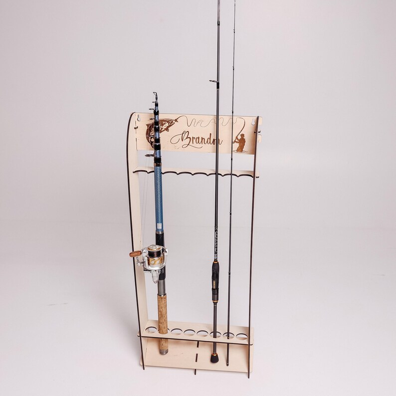 May include: A wooden fishing rod rack with three fishing rods. The rack has a decorative top with the name "Brandon" and a silhouette of a fisherman. The rods are black and blue, and the rack is made of light-colored wood.