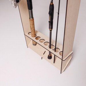 May include: A wooden fishing rod rack with multiple circular openings to hold fishing rods. The rack is made of light-colored wood and has a simple, functional design. Several fishing rods are placed in the rack.