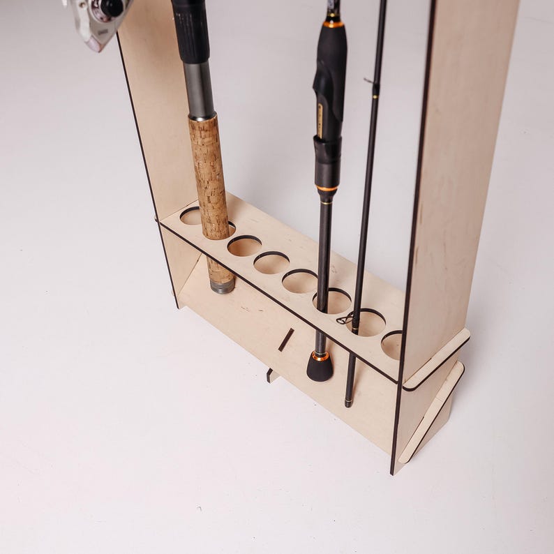 May include: A wooden fishing rod rack with multiple circular openings to hold fishing rods. The rack is made of light-colored wood and has a simple, functional design. Several fishing rods are placed in the rack.