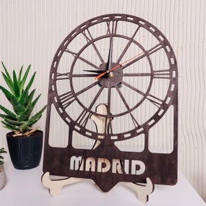 May include: A dark brown wooden clock with Roman numerals and the word "MADRID" cut out at the bottom. The clock has a decorative, open design and is supported by a light-colored wooden stand. The clock hands are black and red.