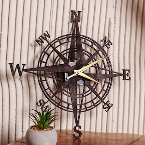 May include: A dark brown compass rose wall clock with gold-colored hands. The clock features directional markers: N, S, E, W, NW, NE, SW, and SE. The clock is made of wood and has a decorative design.