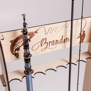 May include: Wooden fishing rod rack with a laser-etched design. Features a fish, a fisherman, and the name "Brandon" in cursive. The rack holds multiple rods and has a light wood finish.