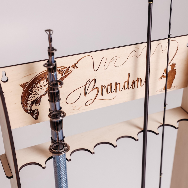 May include: Wooden fishing rod rack with a laser-etched design. Features a fish, a fisherman, and the name "Brandon" in cursive. The rack holds multiple rods and has a light wood finish.
