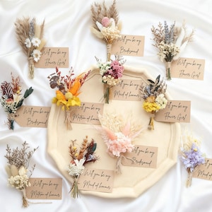 Puede incluir: Un conjunto de ramos de flores secas con etiquetas adjuntas. Las etiquetas dicen "¿Quieres ser mi niña de las flores?", "¿Quieres ser mi dama de honor?" y "¿Quieres ser mi dama de honor?". Los ramos presentan varios colores y texturas, dispuestos sobre una bandeja crema.
