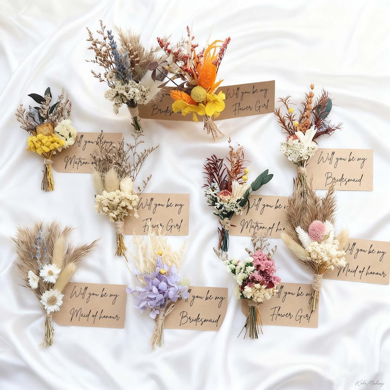 May include: A collection of dried flower bouquets in various colors, including yellow, orange, purple, and white, tied with twine. Each bouquet is paired with a small card that reads "Will you be my..." followed by a wedding party role.