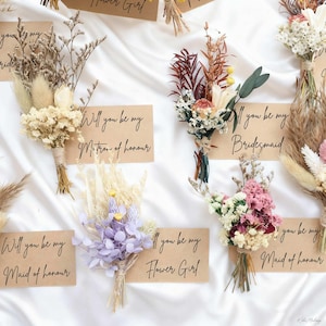 May include: Arrangement of dried flower bouquets with accompanying cards. The cards read "Will you be my Matron of Honor", "Bridesmaid", "Maid of Honor", and "Flower Girl". The bouquets feature various colors and textures.