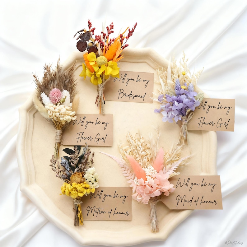 May include: A collection of dried flower bouquets in various colors, including yellow, orange, pink, and purple. Each bouquet is tied with twine and has a small card that reads "Will you be my Bridesmaid", "Flower Girl", "Matron of Honour", or "Maid of Honour".