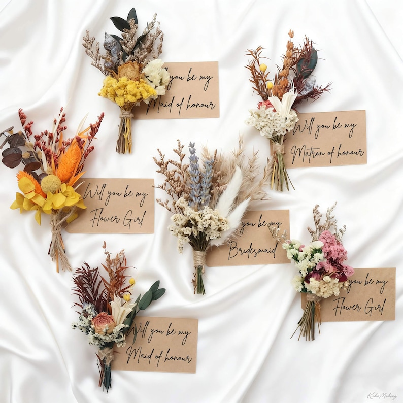 May include: Arrangement of dried flowers in various colors, including yellow, orange, and pink, tied with twine. Each bouquet is paired with a small card that reads "Will you be my..." followed by a wedding party role.