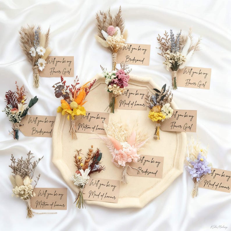 May include: A collection of dried flower bouquets with attached tags. The tags read "Will you be my Flower Girl", "Will you be my Bridesmaid", and "Will you be my Maid of Honour". The bouquets feature various colors and textures, arranged on a cream-colored tray.