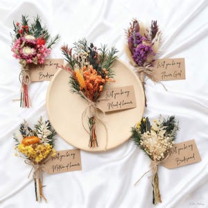 May include: Arrangement of dried flowers in various colors, including pink, orange, yellow, and purple. Each bouquet is tied with twine and includes a tag that reads "Will you be my..." followed by a role such as "Bridesmaid" or "Maid of Honor".