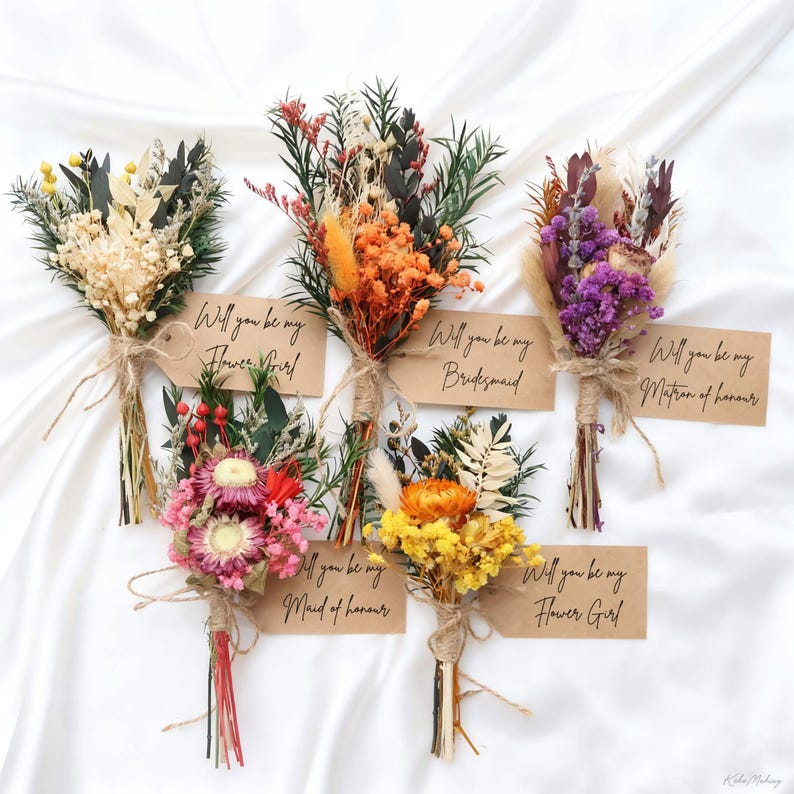 May include: A collection of small, dried flower bouquets in various colors, including orange, yellow, and purple. Each bouquet is tied with twine and has a small tag that reads "Will you be my Flower Girl", "Bridesmaid", "Maid of Honour", or "Matron of Honour".