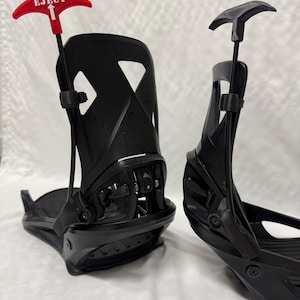 Pode incluir: Um par de fixações de snowboard pretas. Uma das fixações tem uma alavanca vermelha com a palavra "EJECT" impressa. As fixações são projetadas para prender as botas de um snowboarder à prancha. Feitas de plástico e metal duráveis.